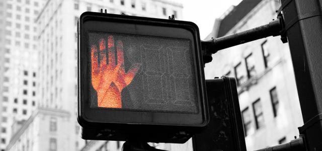 A crosswalk sign in a city. A red hand on the sign is lit up, signaling people to stop